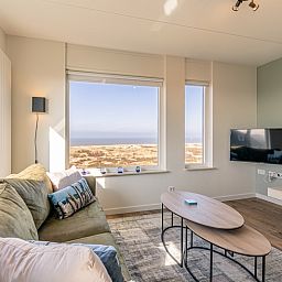 Wohnung Juliana 150 Zeezicht in De Koog Texel, Wohnzimmer mit grossem Fenster und schoener Aussicht auf die Kueste.
