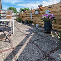 Geraeumige Terrasse am Ferienhaus 2490 in De Koog, Texel, umgeben von einem Holzzaun und bunten Blumen.