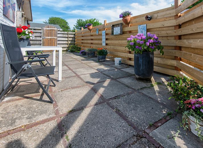 Sonnige Terrasse des Ferienhauses 2490 in De Koog, Texel, mit bequemen Stuehlen und dekorativen Blumenkuebeln.